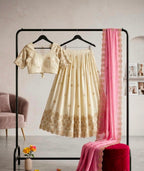 A cream-colored traditional outfit displayed on a black clothing rack, featuring a short-sleeve crop top and a matching pleated skirt with gold floral embroidery and scalloped edges. A pink dupatta with lace trim is draped alongside the outfit, with a red velvet stool and a pink chair visible in the styled room setting.