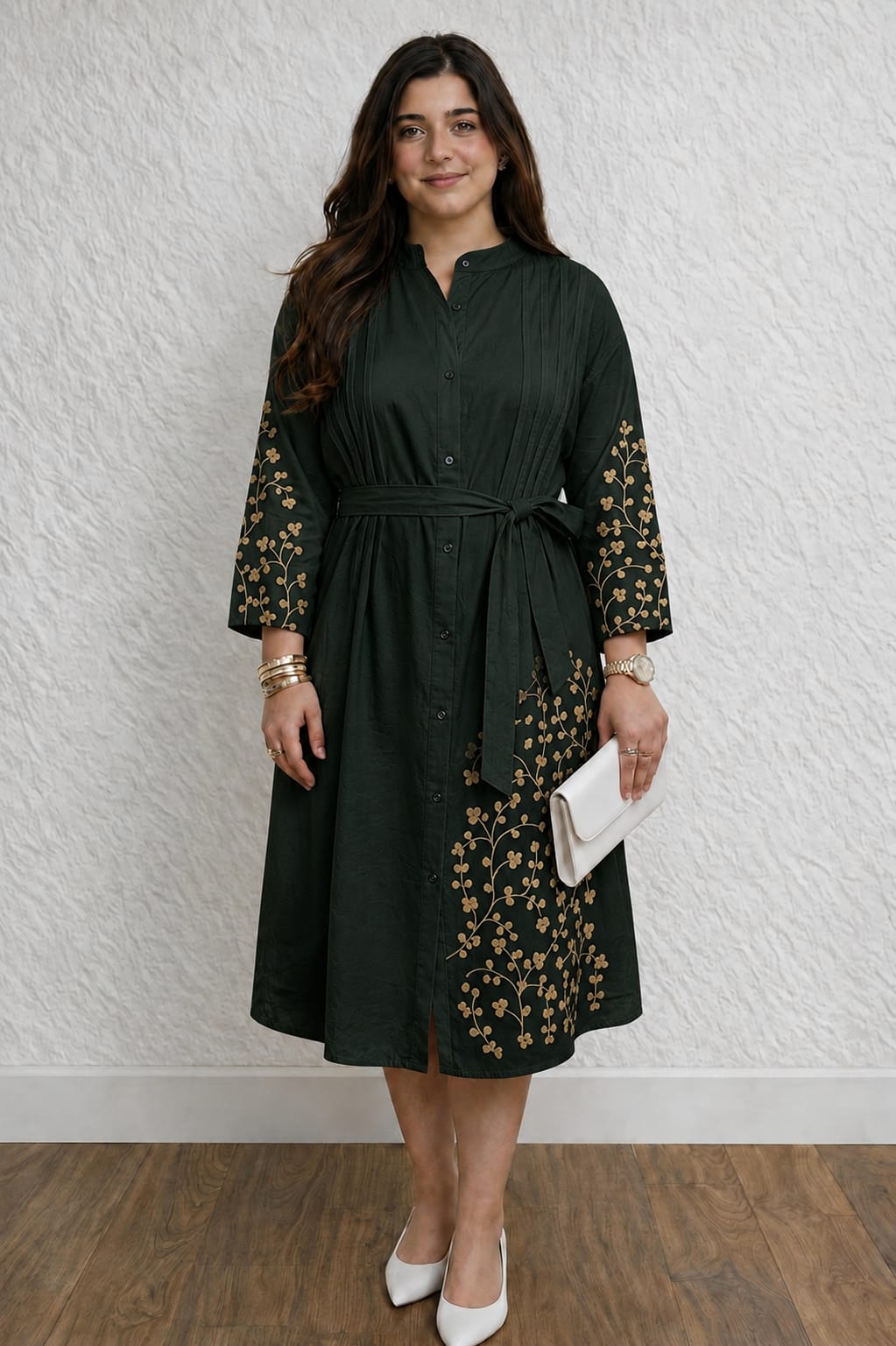 A woman modeling a dark green midi dress with gold floral embroidery on the sleeves and skirt, featuring a tie waist and button front, paired with white heels and holding a white clutch against a textured white wall.