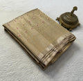 A folded beige silk saree with intricate gold zari embroidery, red dot accents, and a wide striped border, displayed next to a brass incense burner on a textured white fabric background.