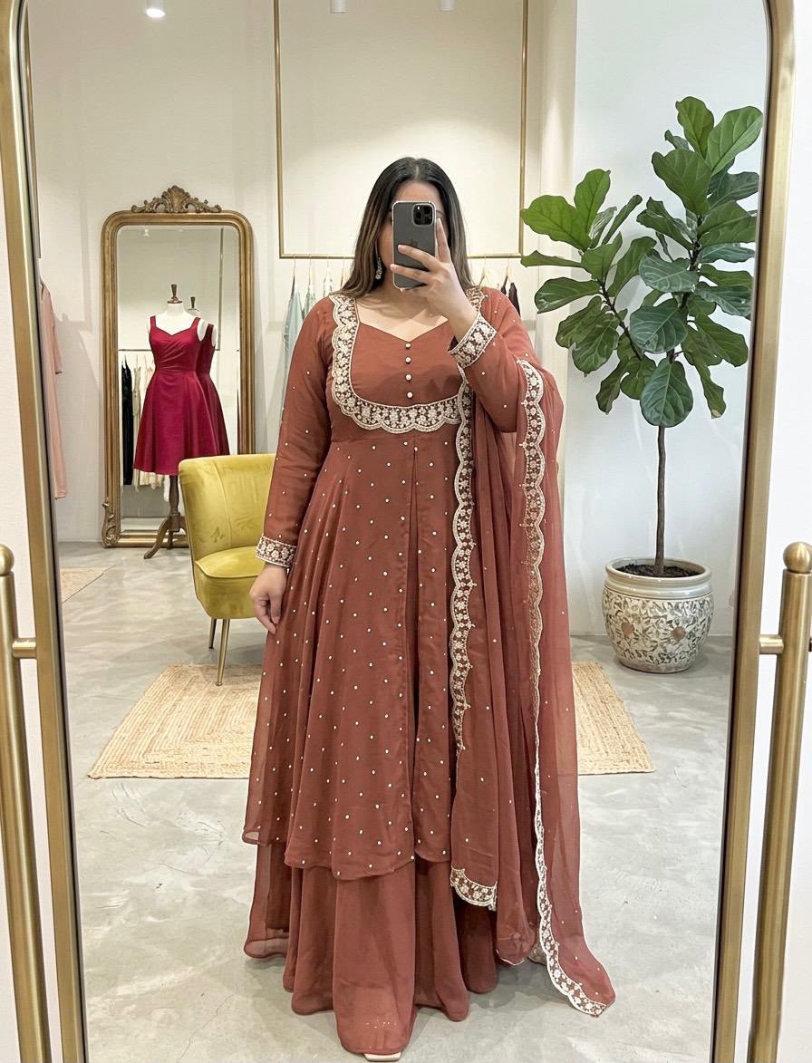 A woman takes a mirror selfie in a boutique wearing a rust-brown Anarkali suit with white embroidery and a matching dupatta. The background shows a yellow chair, a gold-framed mirror, and a potted fiddle-leaf fig plant.