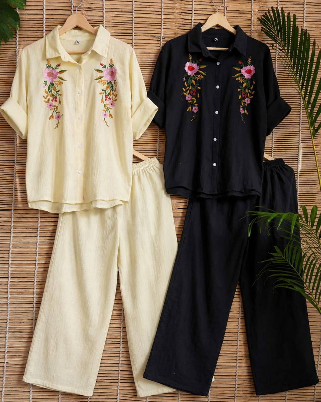 Two matching two-piece linen pajama sets displayed side by side on wooden hangers against a bamboo mat background. The left set is cream-colored with pink floral embroidery on the chest, and the right set is black with matching pink floral embroidery.