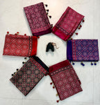 Five folded patterned fabric pieces in red, magenta, purple, black, and maroon are arranged in a circle on a light marble floor, with a small black pouch in the center. Each fabric has ornate circular motifs and pom-pom tassels.