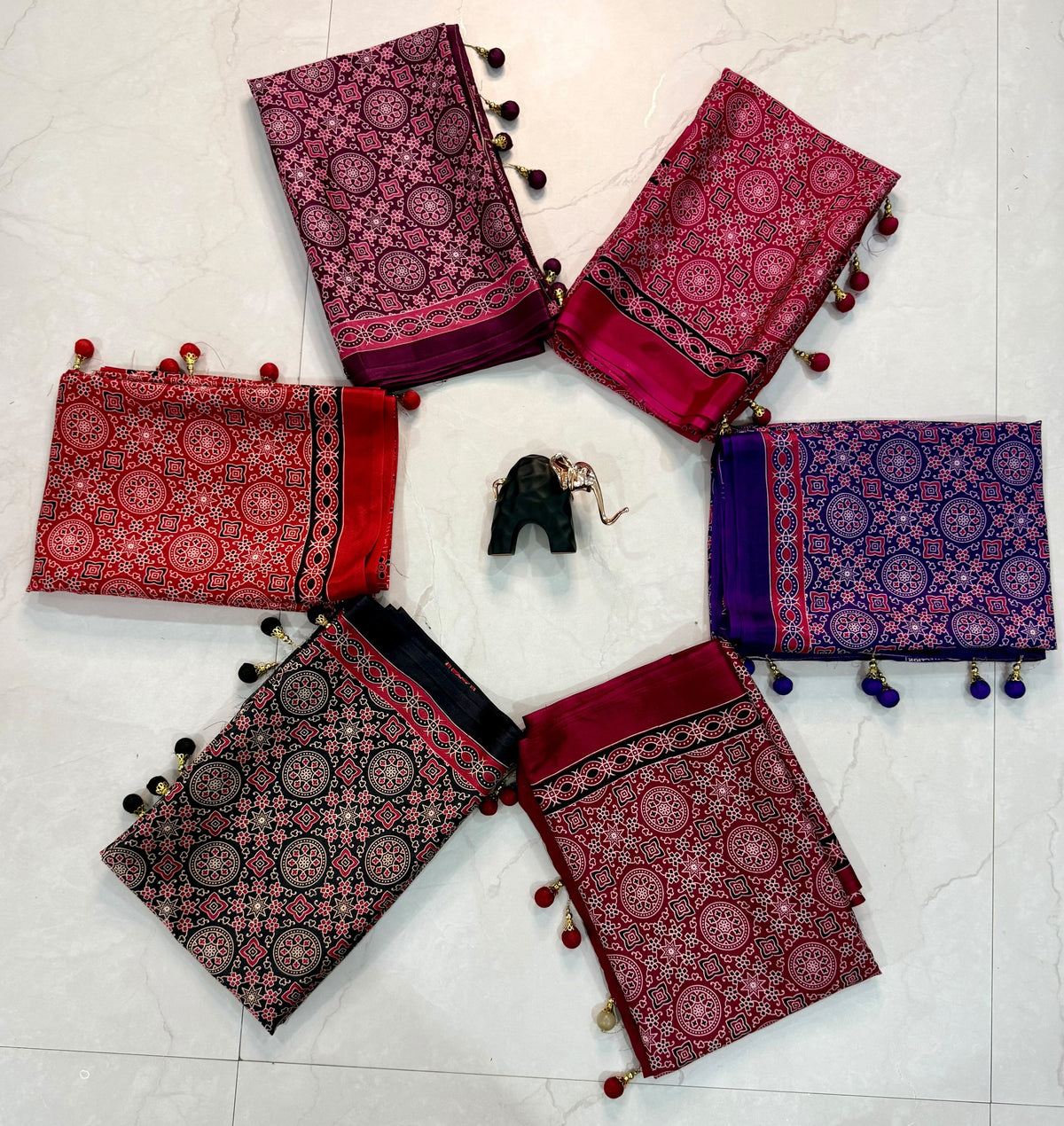 Five folded patterned fabric pieces in red, magenta, purple, black, and maroon are arranged in a circle on a light marble floor, with a small black pouch in the center. Each fabric has ornate circular motifs and pom-pom tassels.