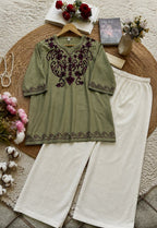 An olive green tunic top with dark purple embroidery, paired with off-white wide-leg pants, arranged in a styled flat-lay on a woven mat with flowers, books, and a book as props.