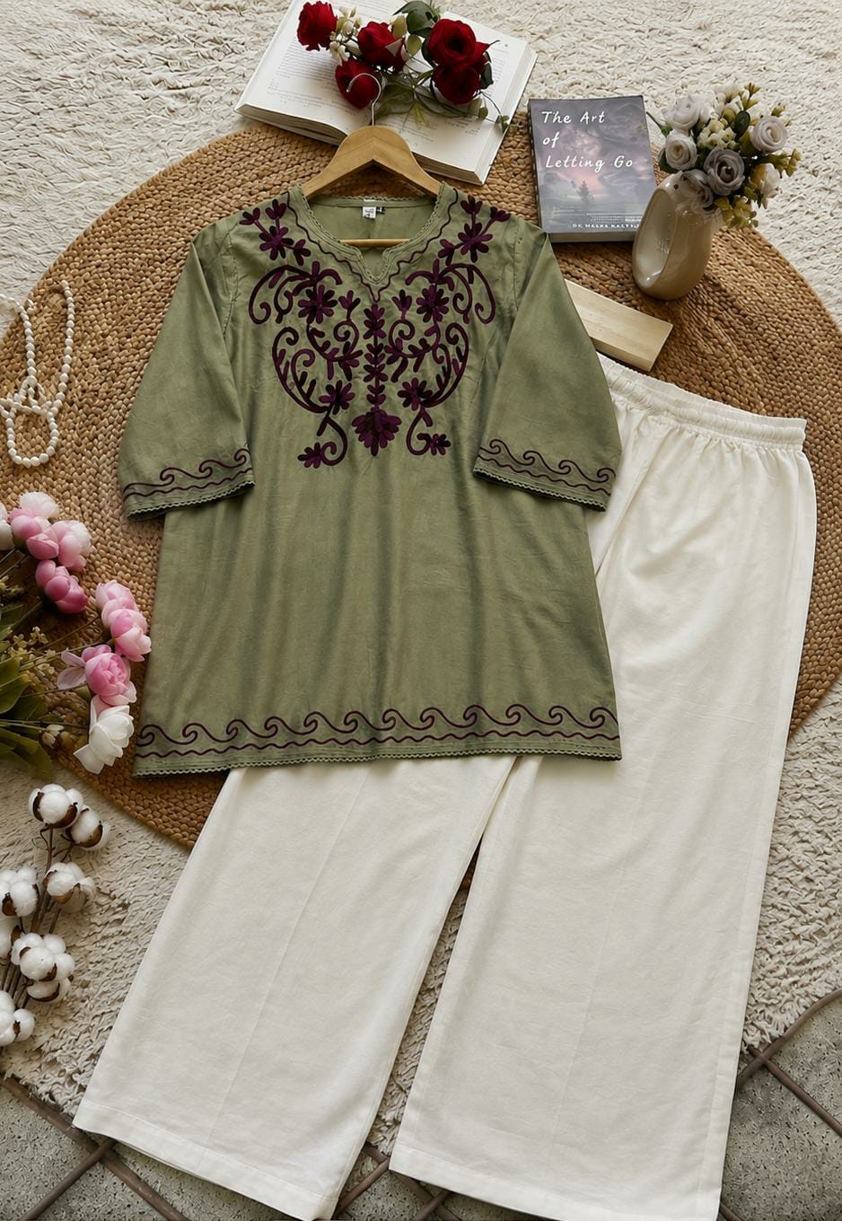 An olive green tunic top with dark purple embroidery, paired with off-white wide-leg pants, arranged in a styled flat-lay on a woven mat with flowers, books, and a book as props.