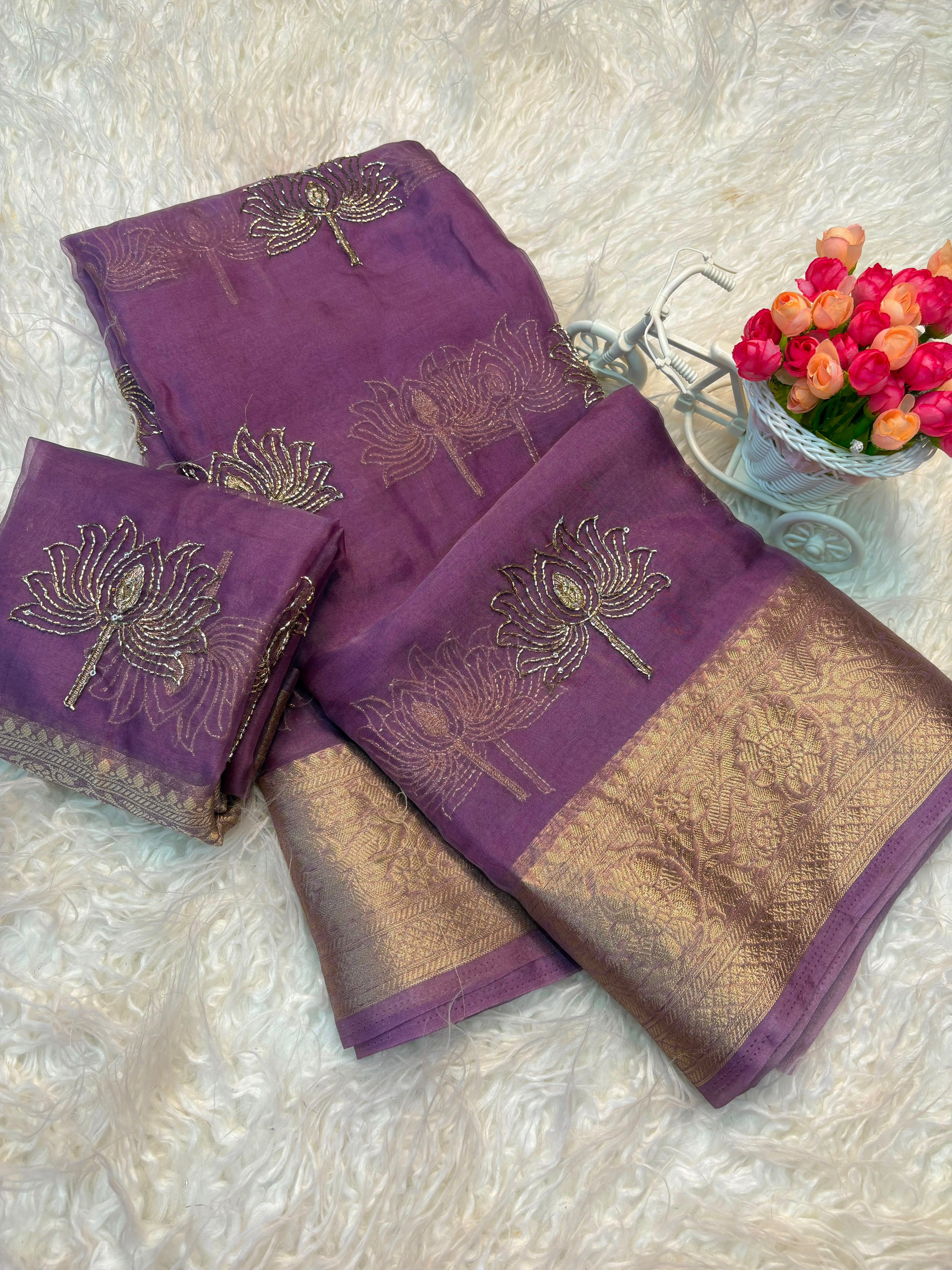 Purple silk saree with gold zari floral embroidery and a wide gold brocade border, displayed on a white faux fur surface next to a small decorative flower pot.
