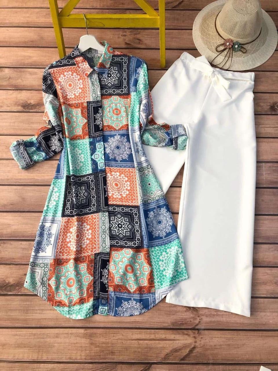 A women's long-sleeve dress with a colorful patchwork print in teal, orange, navy, and white, styled next to a solid white long-sleeve top and a straw hat on a wooden plank background.