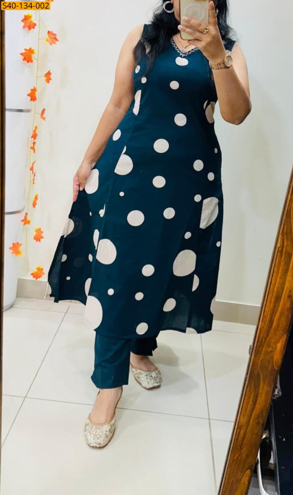 A woman takes a mirror selfie wearing a sleeveless dark teal blue A-line kurta with large white polka dots, paired with matching teal pants and beige embellished flat shoes.