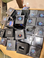 A pile of boxed smartwatches in their original black retail packaging, with one watch and a USB cable visible outside the boxes on top.