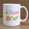 White mug with 'Sweet' in pink and floral designs on a wooden surface.