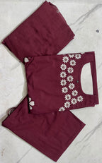 A folded three-piece women's salwar kameez set in maroon, featuring a kurta with white floral embroidery along the sleeve, paired with matching trousers and a dupatta, laid flat on a light marble surface.