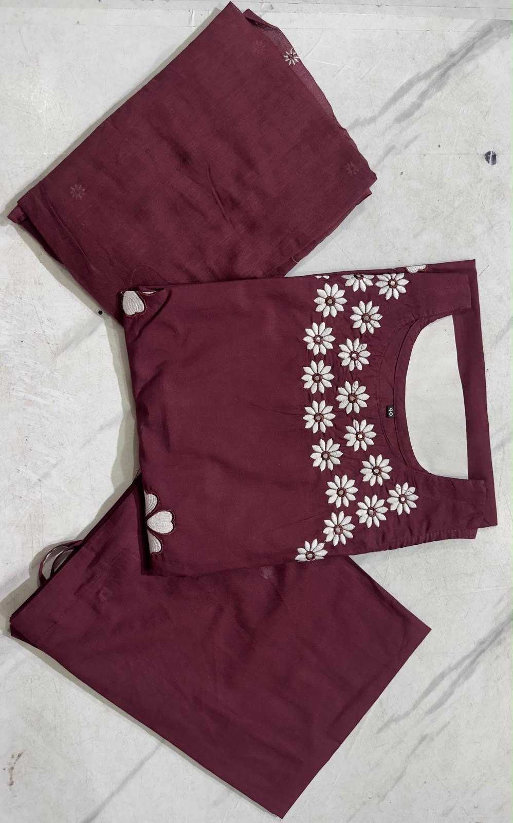 A folded three-piece women's salwar kameez set in maroon, featuring a kurta with white floral embroidery along the sleeve, paired with matching trousers and a dupatta, laid flat on a light marble surface.