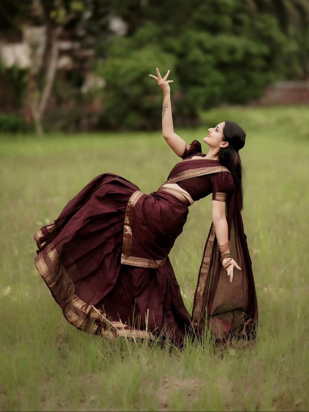 A woman in a dark maroon and gold saree poses dynamically in a grassy field, looking upwards with one arm raised. The background shows a soft-focus green landscape with trees.