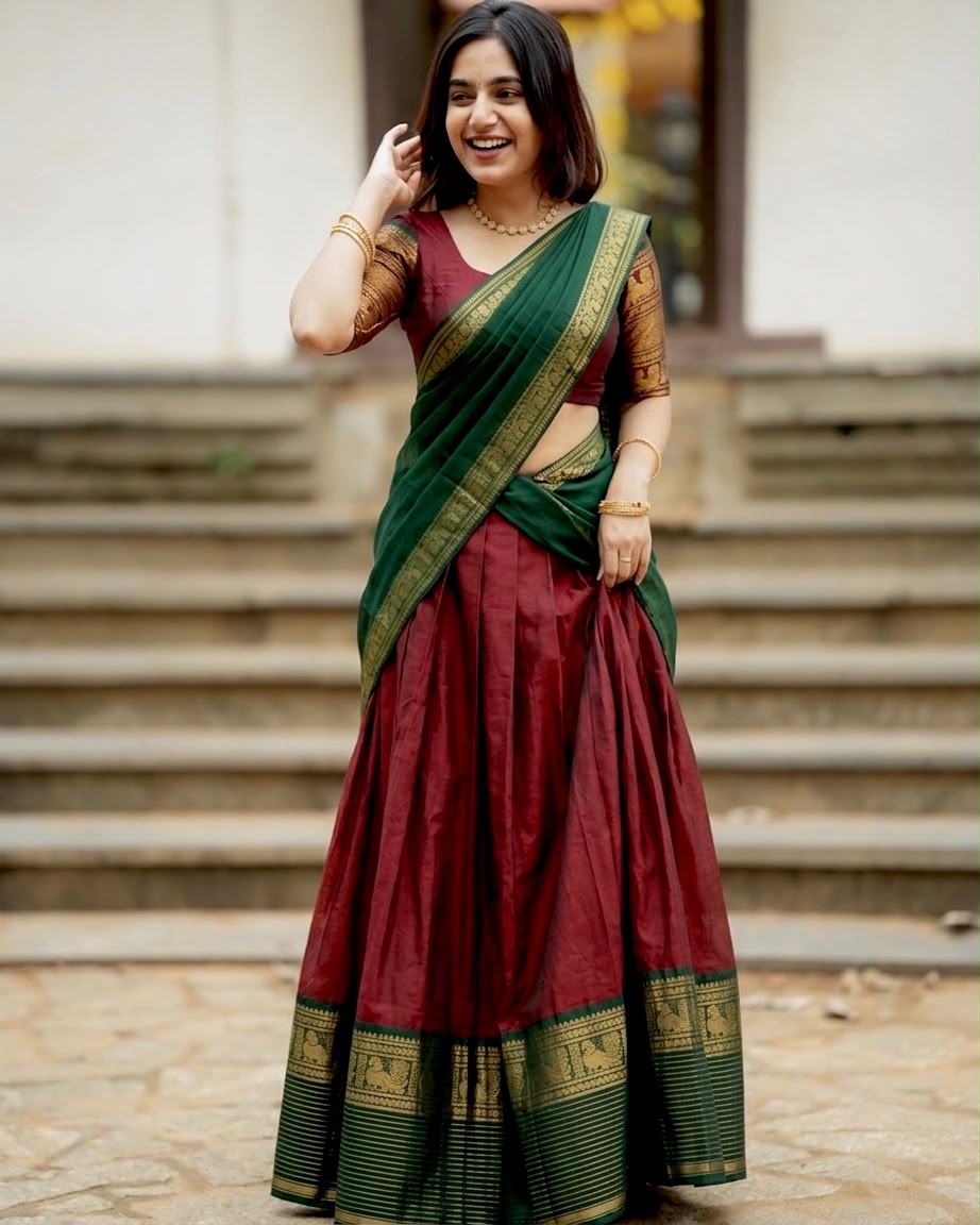 A woman wearing a traditional Indian lehenga choli saree in deep maroon and forest green with gold zari borders, standing in front of outdoor stone steps.