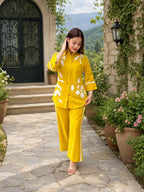 A woman wearing a bright yellow two-piece pajama set with white floral embroidery walks barefoot on a stone path in front of a stone building with potted plants.
