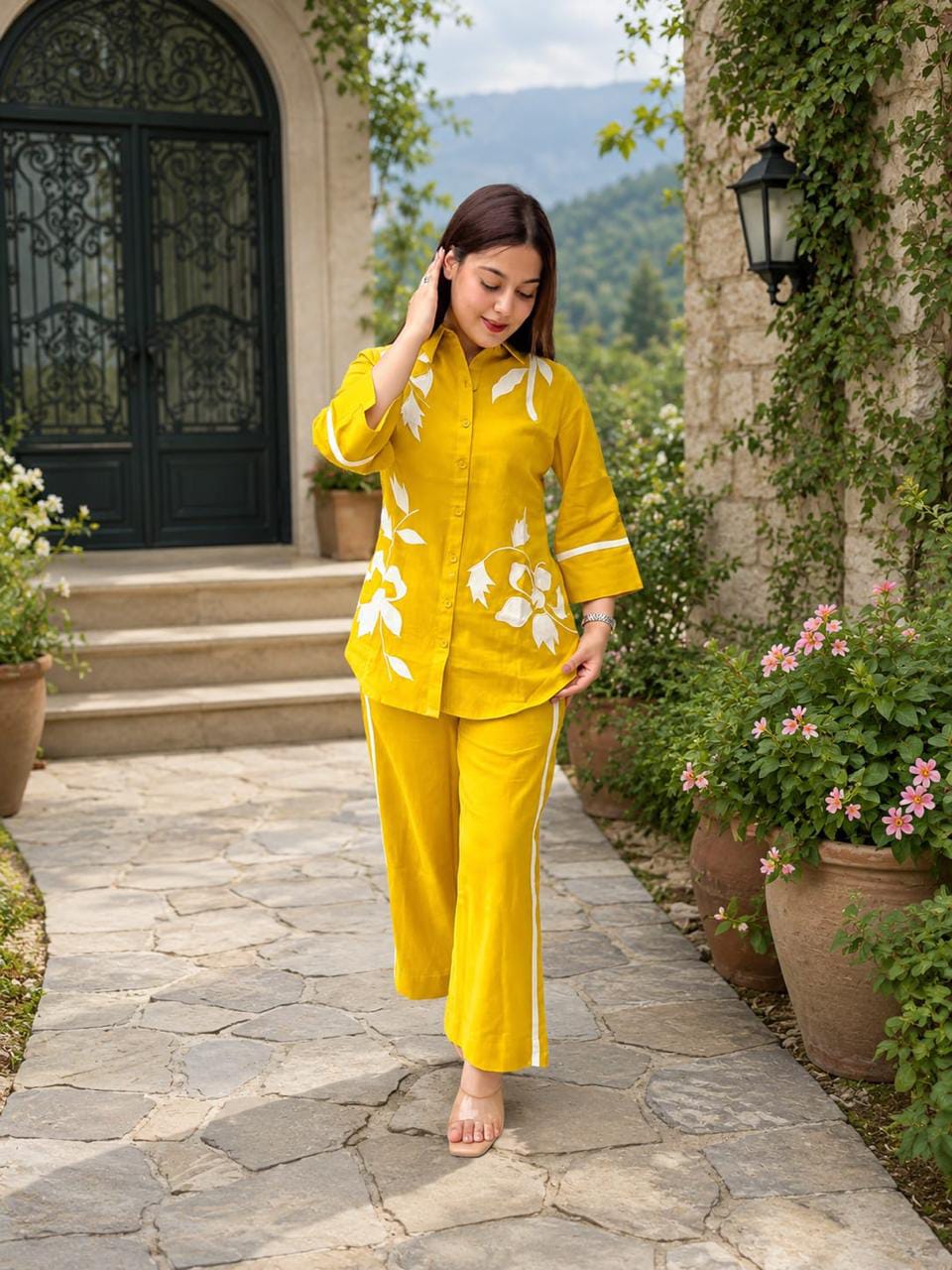 A woman wearing a bright yellow two-piece pajama set with white floral embroidery walks barefoot on a stone path in front of a stone building with potted plants.