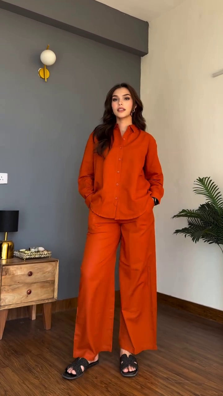 Woman in an orange outfit standing in a room with a gray wall and wooden furniture.