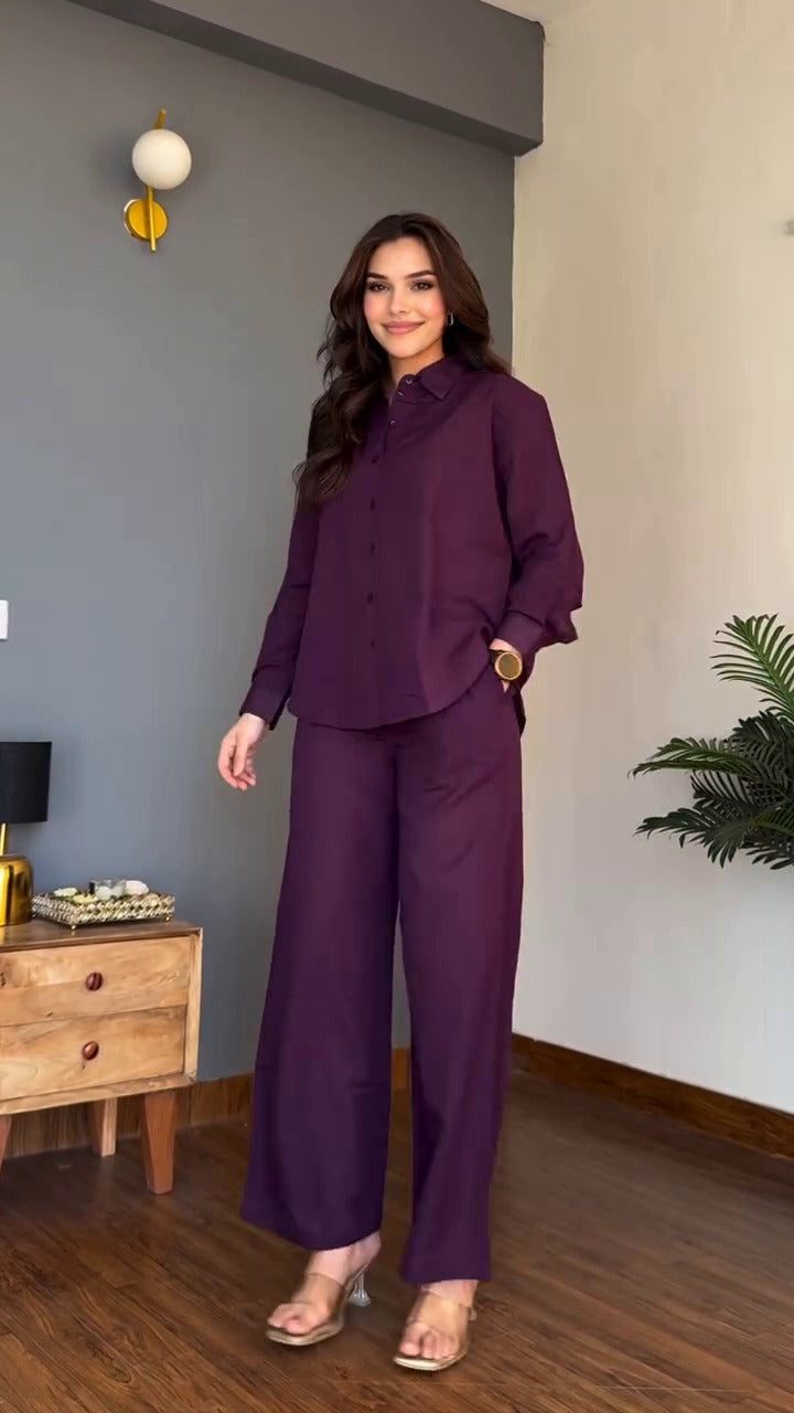 Woman in a purple outfit standing in a room with a gray wall and wooden furniture.
