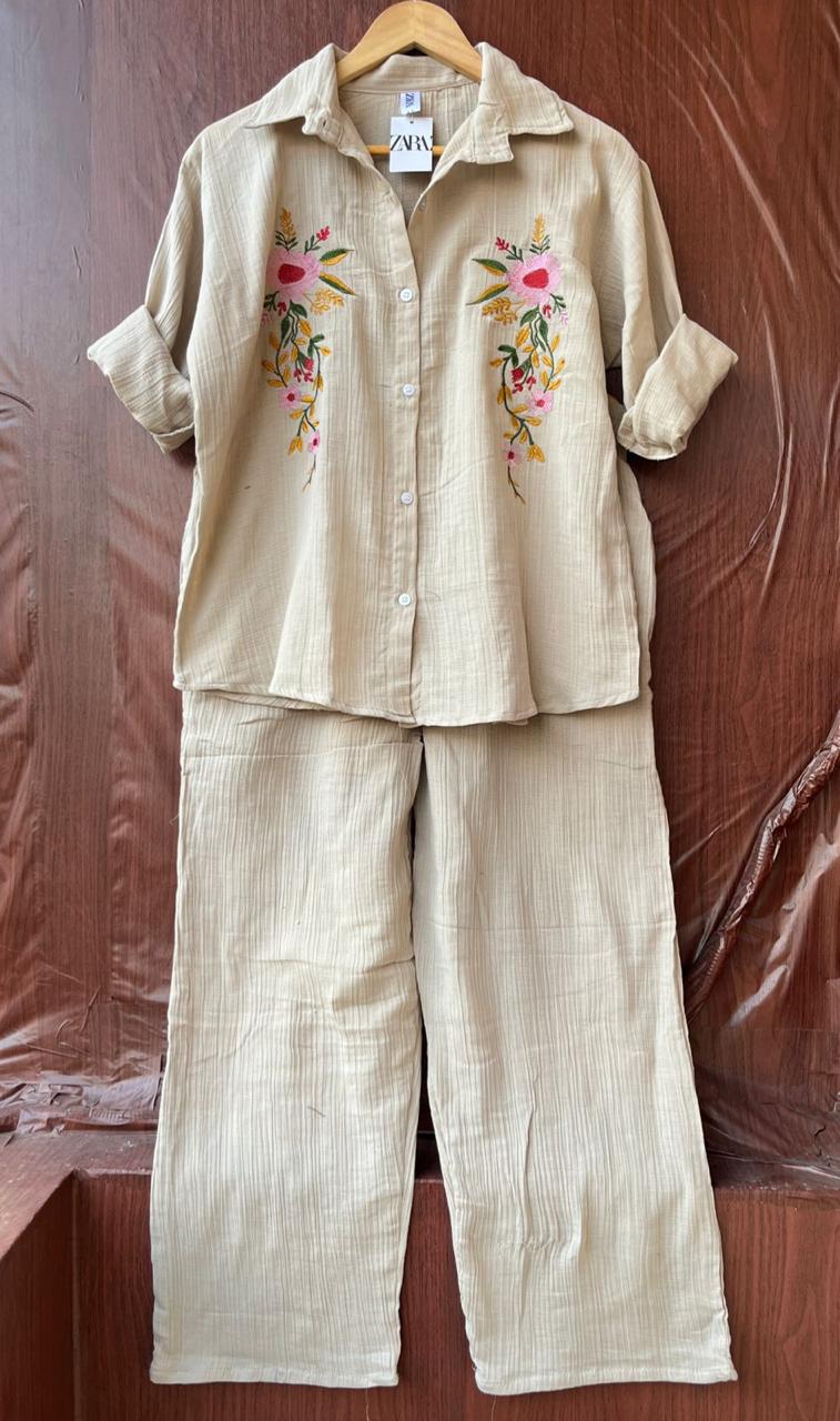 A two-piece linen pajama set in beige, featuring a short-sleeve button-up shirt with colorful floral embroidery on the chest and matching wide-leg pants, hanging on a wooden hanger against a brown textured backdrop.