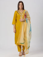 A woman modeling a mustard yellow traditional kurta set with matching pants and a sheer printed dupatta, photographed against a light gray studio background.