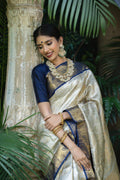 Woman in traditional attire with a blue blouse and white saree, standing among green plants.