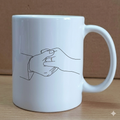 White mug with a line drawing of two hands holding each other on a wooden surface.
