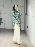 A woman takes a mirror selfie wearing a long-sleeve, high-neck blouse with a green, blue, and orange abstract pattern, paired with cream-colored wide-leg trousers and beige heeled sandals.