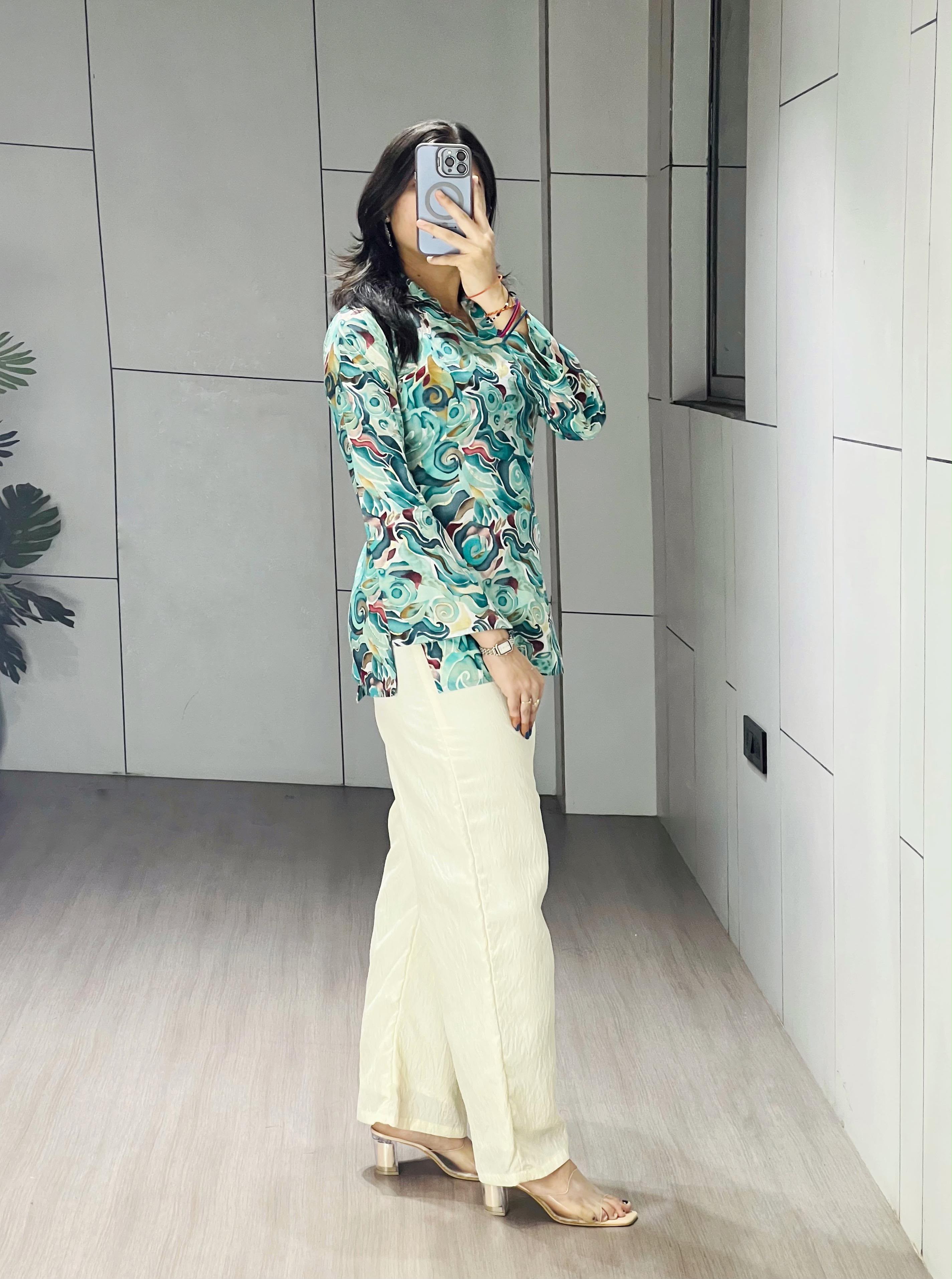 A woman takes a mirror selfie wearing a long-sleeve, high-neck blouse with a green, blue, and orange abstract pattern, paired with cream-colored wide-leg trousers and beige heeled sandals.