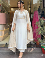 Woman in a white traditional outfit with gold embroidery standing in front of a store entrance.