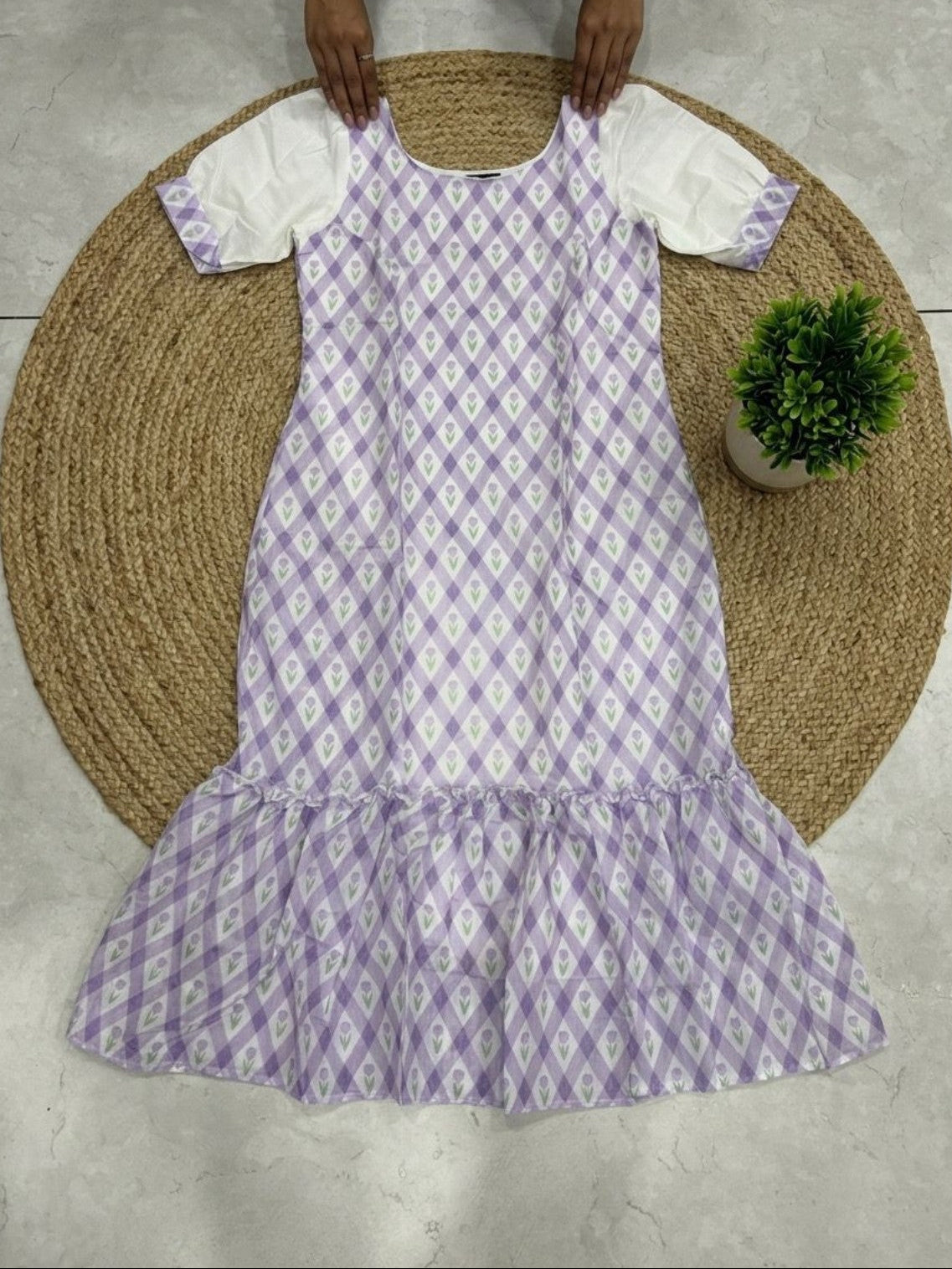 A sleeveless babydoll dress with a purple and white ikat pattern, featuring white puff sleeves, held up by a person's hands over a round woven jute mat.