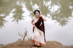 A woman wearing a traditional Indian saree stands outdoors near water, holding the fabric. The saree has a deep maroon upper drape and a light cream lower skirt with a decorative border, paired with a cream blouse.