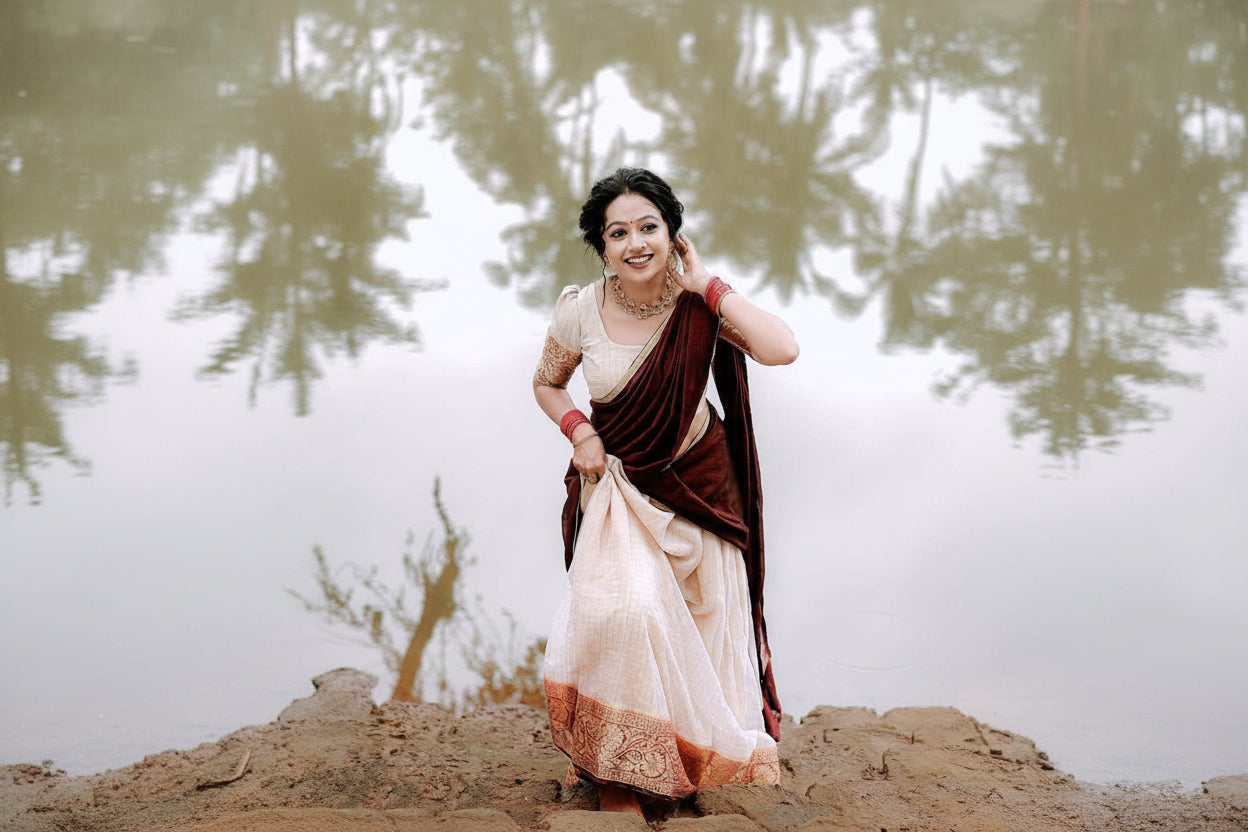 A woman wearing a traditional Indian saree stands outdoors near water, holding the fabric. The saree has a deep maroon upper drape and a light cream lower skirt with a decorative border, paired with a cream blouse.