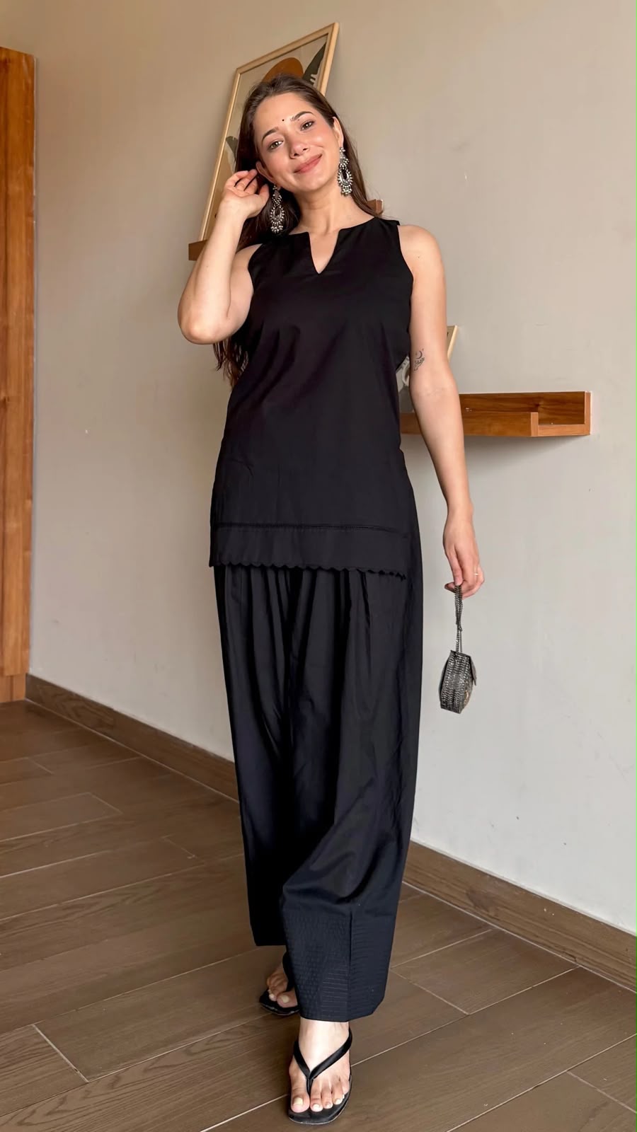 Woman wearing a black sleeveless top and wide-leg pants, holding a handbag indoors.