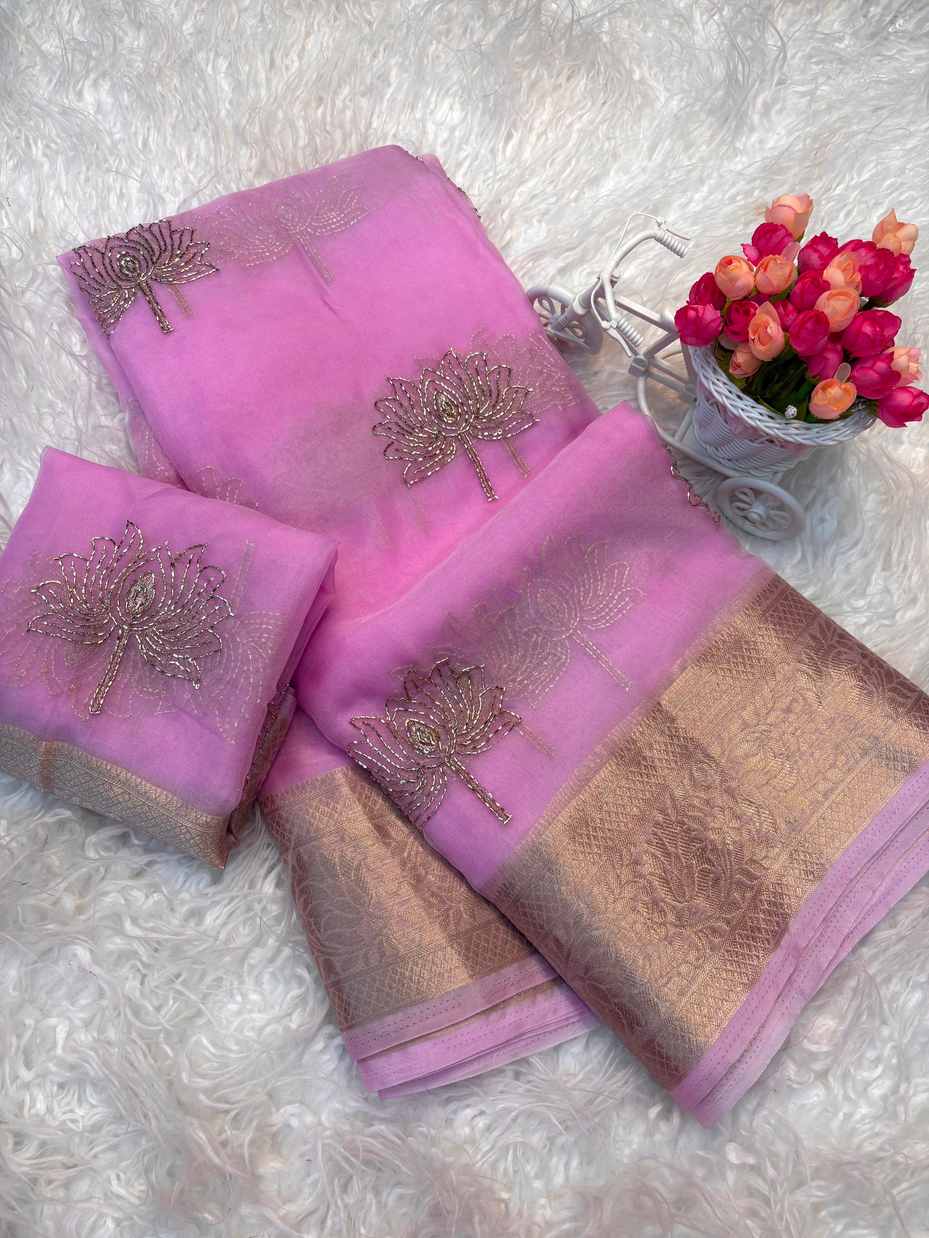 A pink traditional fabric set with metallic gold embroidered motifs and a gold patterned border, arranged on a white faux fur surface next to a small decorative vase of pink flowers.