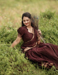 A woman wearing a dark maroon traditional dress with gold embroidery sits in a grassy field, holding a peacock feather bundle.