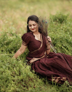 A woman wearing a dark maroon traditional dress with gold embroidery sits in a grassy field, holding a peacock feather bundle.