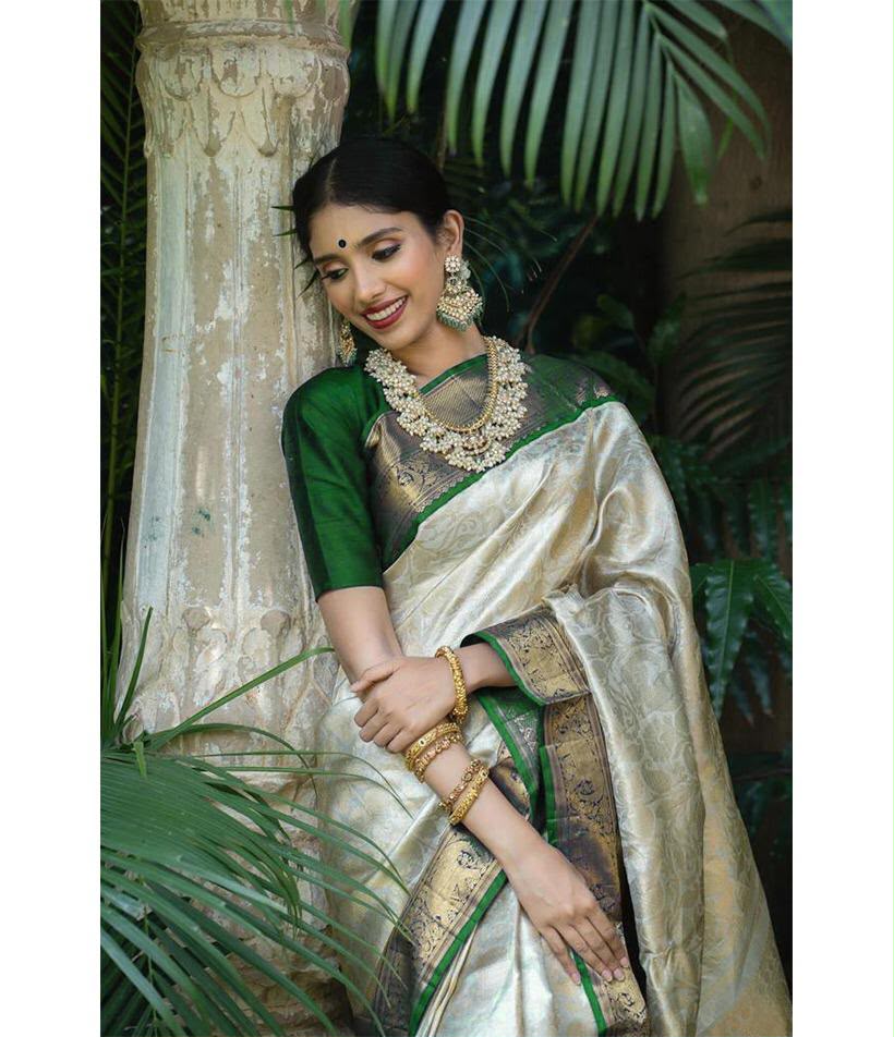 Woman in a traditional saree with green and gold accents, standing among plants.