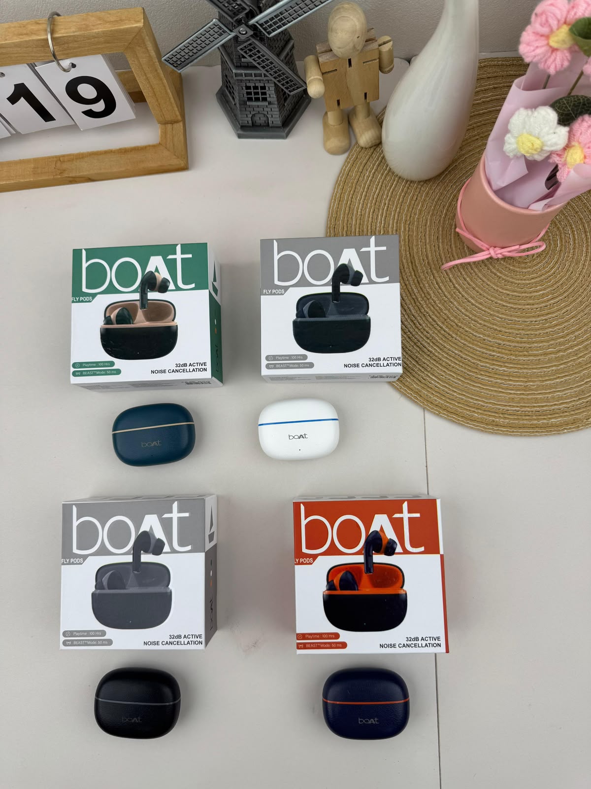 Four pairs of boAt wireless earbuds in their original retail packaging, arranged on a light surface with decorative props including a wooden calendar, a small windmill, a vase, and a pink flower pot.