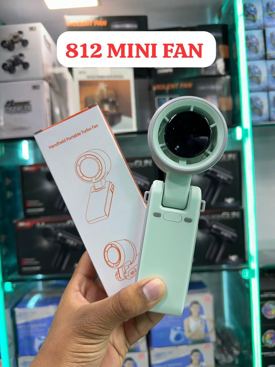 A hand holds a mint green handheld portable tube fan next to its white packaging box in a retail store setting. A white text bubble at the top reads '812 MINI FAN'.