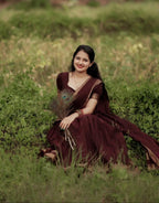 A woman wearing a dark maroon sari sits in a grassy outdoor field, holding a peacock feather. The sari has gold zari embroidery along the border.
