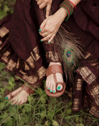 A person outdoors wearing brown leather thong sandals with turquoise painted toenails, a dark brown and gold patterned skirt, and green/red bangles, seated on grass.