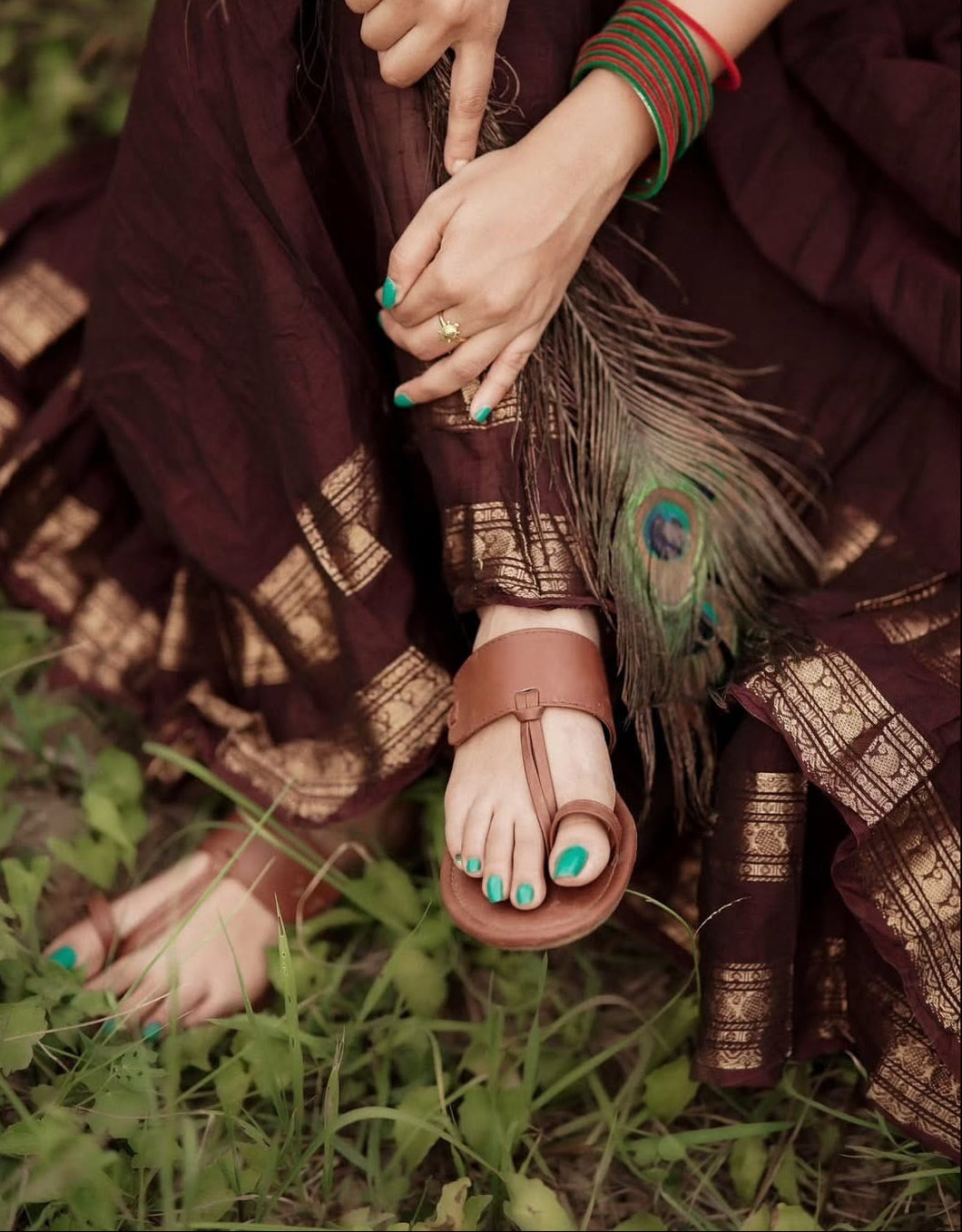 A person outdoors wearing brown leather thong sandals with turquoise painted toenails, a dark brown and gold patterned skirt, and green/red bangles, seated on grass.