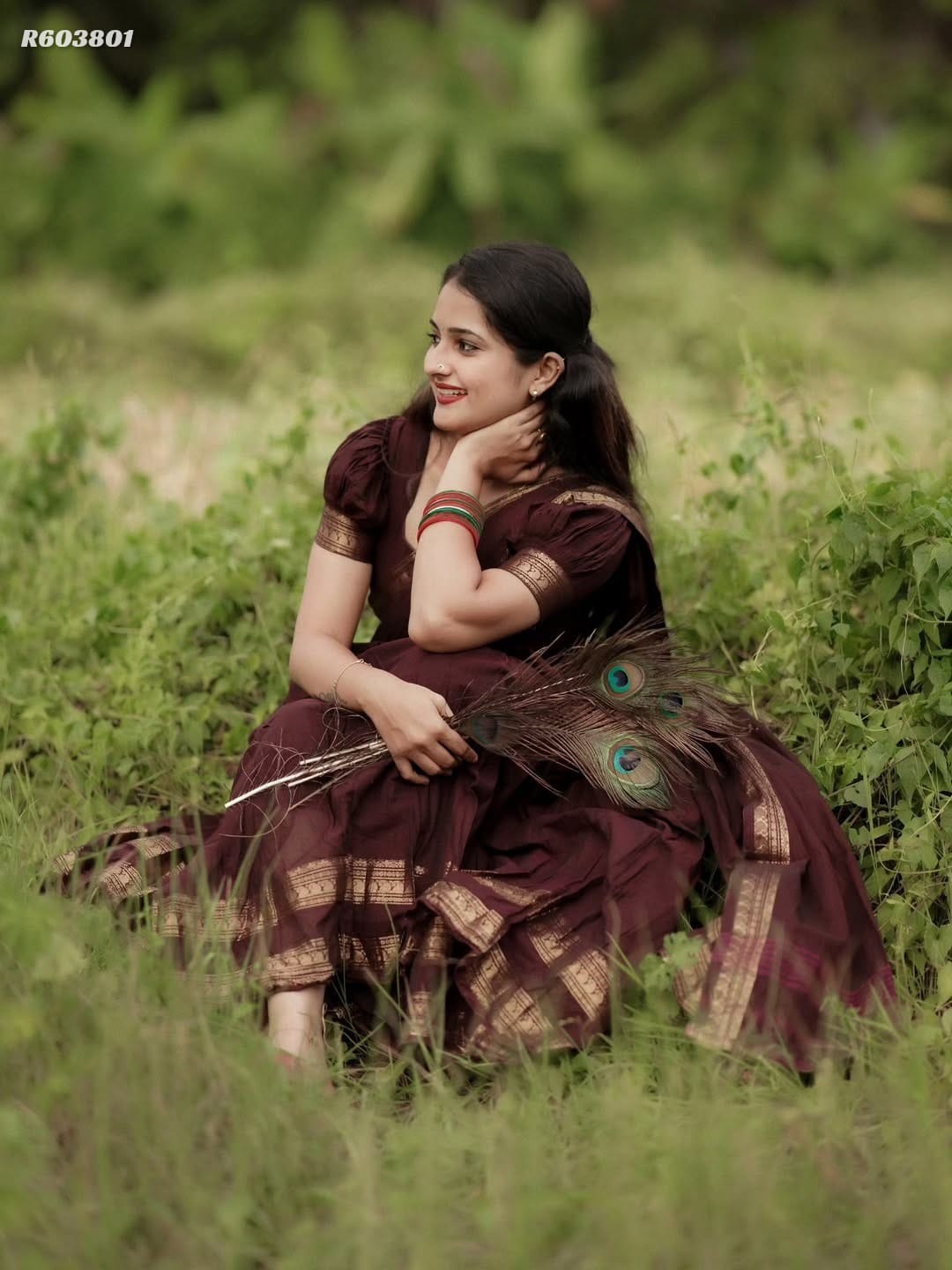 A woman wearing a dark maroon traditional dress with gold patterns sits in a grassy outdoor field, holding peacock feather branches.