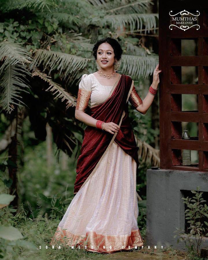 A woman wearing a traditional Indian saree stands outdoors next to a wooden structure. The saree has a deep maroon-red drape over a light beige skirt with gold zari embroidery.