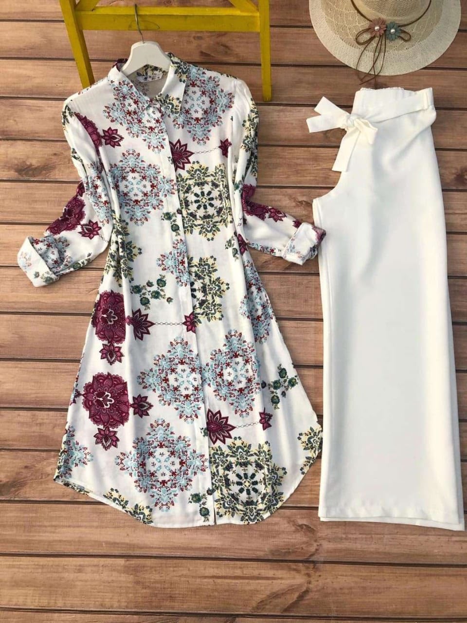 A women's two-piece outfit laid flat on a wooden surface, featuring a long-sleeve button-front dress with a white base and multicolor floral and medallion print, paired with a matching solid white high-waisted skirt or long shorts with a tie waist. A straw hat and yellow stool are partially visible as styling props.