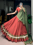 A woman takes a mirror selfie wearing a traditional Indian lehenga choli set. The outfit features a rust-red flared skirt (lehenga) with a green and gold border, a matching rust-red blouse with green and gold trim, and a sage green dupatta with gold motifs.