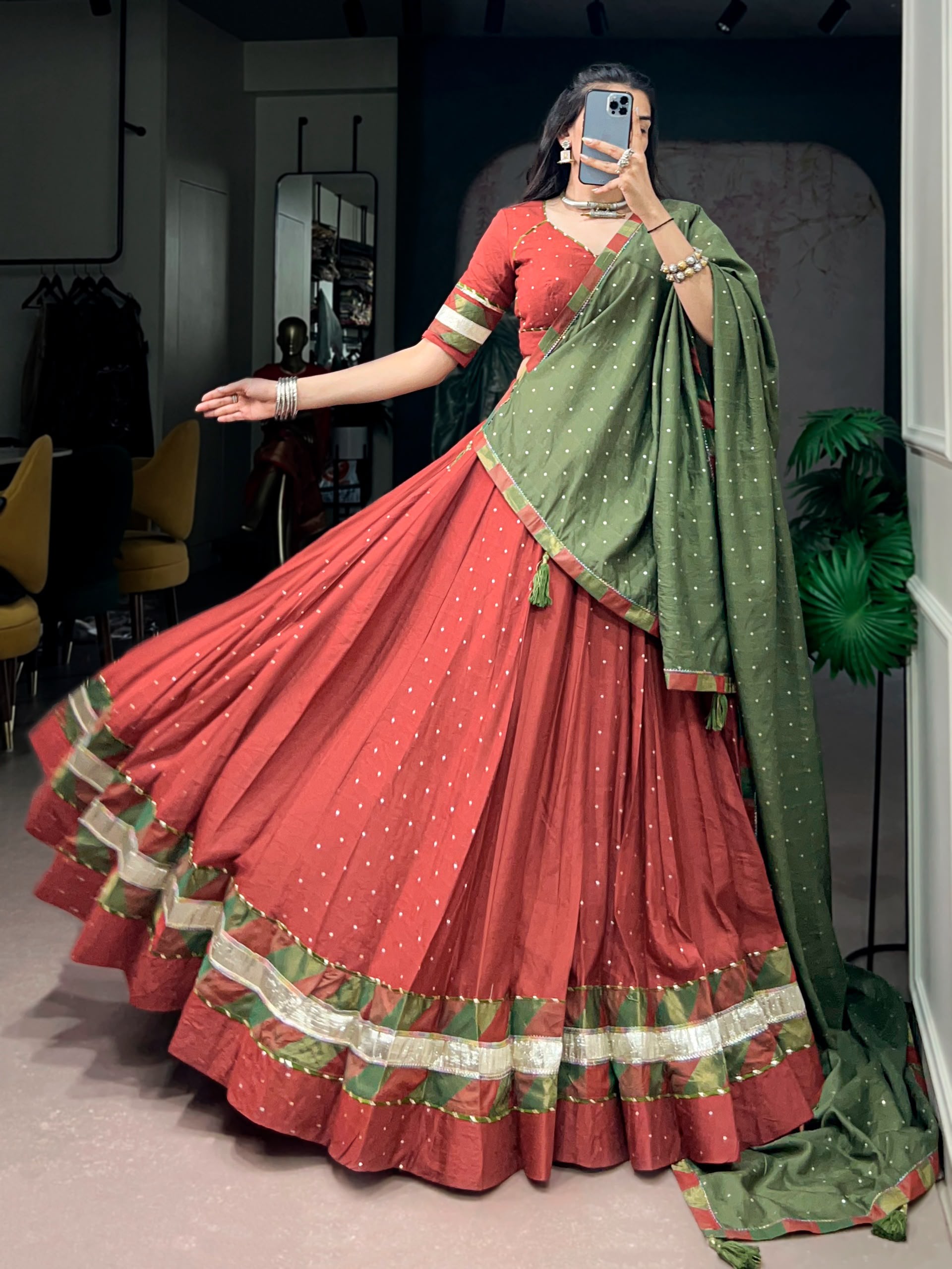 A woman takes a mirror selfie wearing a traditional Indian lehenga choli set. The outfit features a rust-red flared skirt (lehenga) with a green and gold border, a matching rust-red blouse with green and gold trim, and a sage green dupatta with gold motifs.