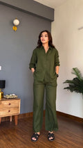 A woman modeling an olive green two-piece pajama set consisting of a long-sleeve button-up shirt and matching wide-leg pants, standing in a modern room with a gray accent wall, wooden floor, and a plant.