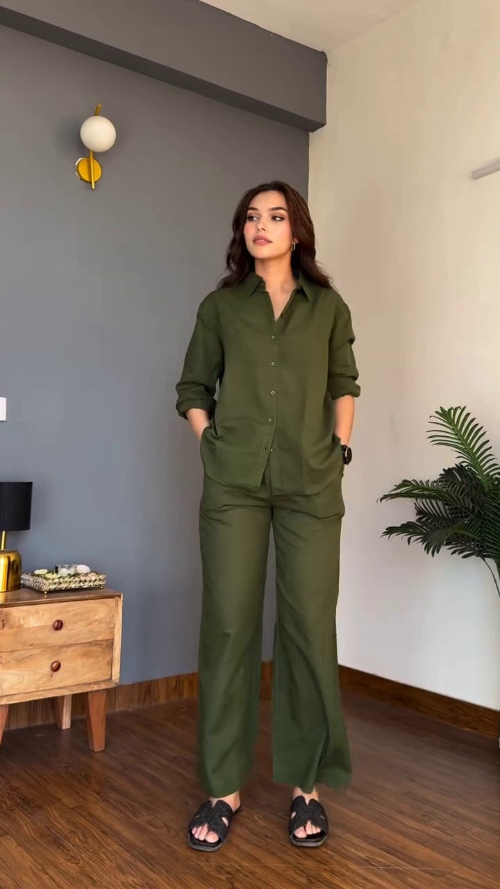 A woman modeling an olive green two-piece pajama set consisting of a long-sleeve button-up shirt and matching wide-leg pants, standing in a modern room with a gray accent wall, wooden floor, and a plant.