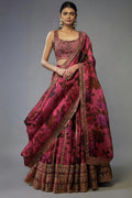 A model wearing a traditional Indian lehenga choli set in a rich magenta pink color with dark floral patterns and heavy gold embroidery, set against a plain gray studio background. A white label with 'D.